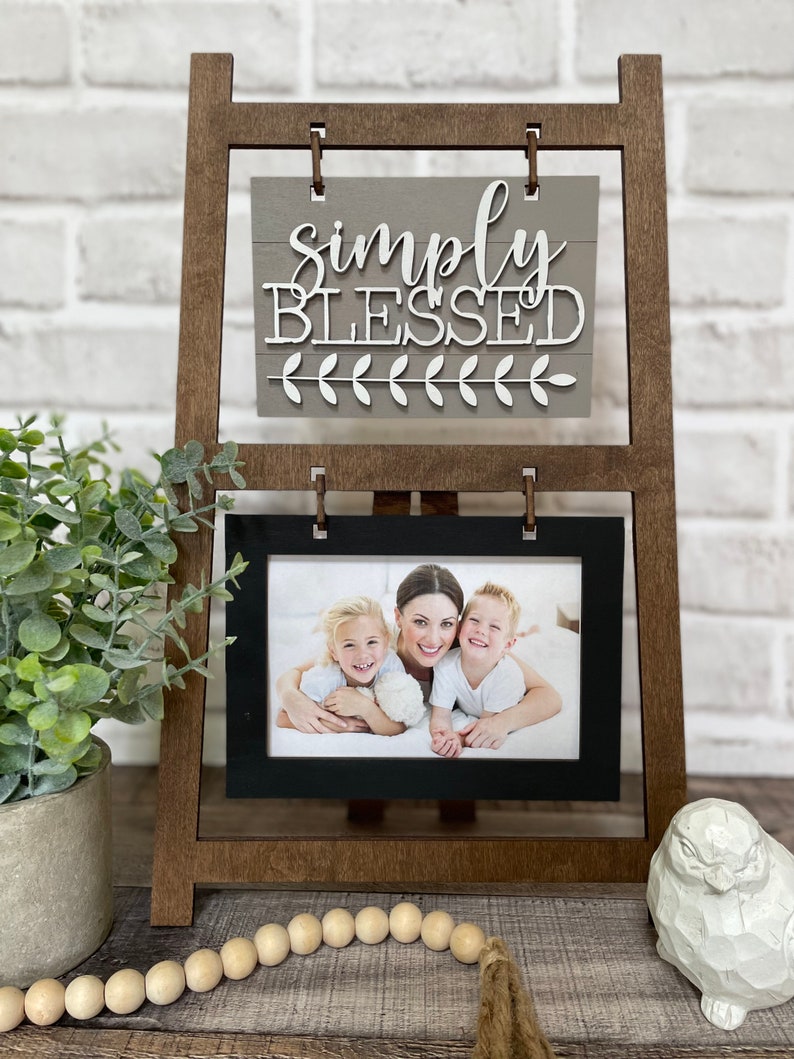 May include: A wooden easel with a gray sign that says "simply blessed" and a black frame with a photo of a mother and her two children. The easel is on a wooden surface with a string of wooden beads and a white owl figurine.