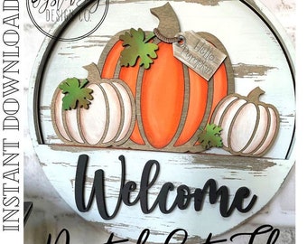 May include: A round wooden sign with a distressed white paint finish. The sign features a layered design of three pumpkins with a tag that reads "Hello Pumpkin". The word "Welcome" is written in black lettering below the pumpkins.