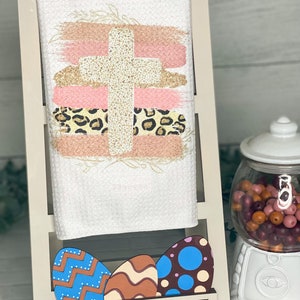 Op de afbeelding: Witte handdoek met een gouden kruis en roze en bruine penseelstreken, een luipaardprint en een bloemenmotief. Op de voorgrond liggen drie houten paaseieren met verschillende patronen.