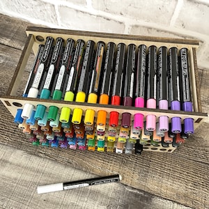 May include: A wooden organizer with rows of colorful acrylic paint pens. The pens are labeled "TOOLI-ART" and have black caps. The organizer has a wooden frame and is designed to hold the pens upright.
