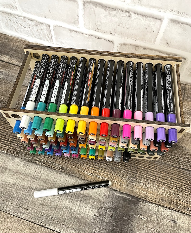 May include: A wooden organizer with rows of colorful acrylic paint pens. The pens are labeled "TOOLI-ART" and have black caps. The organizer has a wooden frame and is designed to hold the pens upright.