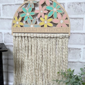 以下が含まれることがあります： 花柄のデザインと「Live by grace not perfection」というテキストが書かれた木製の半円形の壁掛け。壁掛けには白い糸のフリンジが付いている。