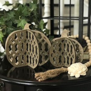 May include: Three wooden pumpkin decorations with a lattice design, perfect for fall. The pumpkins are various sizes and sit on a dark surface with a decorative tassel and a white flower. A string of wooden beads is also present.