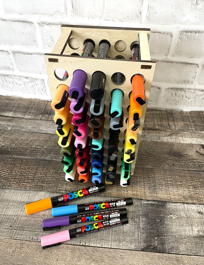 May include: A wooden organizer with 36 holes for storing markers. The organizer is filled with colorful markers, including black, orange, yellow, green, blue, purple, pink, and brown. There are also five markers lying on a wooden surface below the organizer. The markers are labeled "POSCA" in black text.