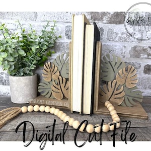Puede incluir: Soportes para libros de madera con diseño de hojas de monstera en tonos verdes y beige. Dos libros se encuentran entre los sujetalibros. Una guirnalda de cuentas de madera y una planta en maceta están en primer plano. El texto "Digital Cut File" está en la parte inferior.
