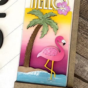 May include: A wooden sign with the word "HELLO" in white letters with a yellow outline. The sign features a pink flamingo standing in blue water with a palm tree and a pink hibiscus flower.