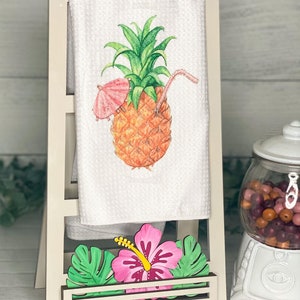 Könnte beinhalten: Ein weißer Handtuchhalter mit einem weißen Handtuch mit einem Ananas-Motiv. Die Ananas hat einen rosa Sonnenschirm und einen Strohhalm. Der Halter hat ein Holzregal mit einer rosa Hibiskusblüte und grünen Blättern.
