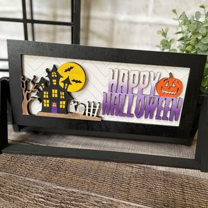 May include: A black framed Halloween decoration with a haunted house, bats, and a pumpkin. The words "HAPPY HALLOWEEN" are displayed in purple and white. The background has a white and gray chevron pattern.