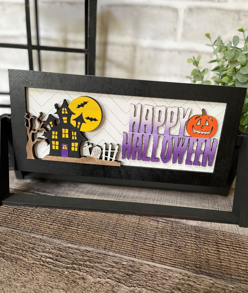 May include: A black framed Halloween decoration with a haunted house, bats, and a pumpkin. The words "HAPPY HALLOWEEN" are displayed in purple and white. The background has a white and gray chevron pattern.