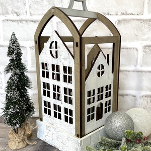 May include: A white wooden house-shaped lantern with a brown wooden frame and a silver metal handle. The lantern has a distressed white paint finish and features cut-out windows. The lantern is surrounded by greenery and silver ornaments.