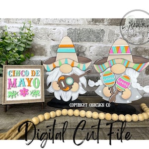 Pode incluir: Dois gnomos de madeira vestidos com sombreros coloridos e segurando instrumentos musicais. O gnomo à esquerda está segurando um violão e o gnomo à direita está segurando maracas. Os gnomos estão em pé em bases pretas e estão em frente a uma placa de madeira que diz "Cinco de Mayo".