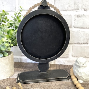 Könnte beinhalten: Rundes Schild aus schwarzem Holz mit einer abgenutzten Oberfläche, das oben einen Perlenbesatz hat. Das Schild steht auf einem schwarzen Ständer mit einem dekorativen Sockel.