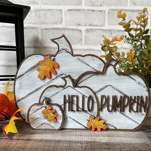 May include: White wooden pumpkin sign with the text "HELLO PUMPKIN" and orange fall leaves. The sign is layered with three pumpkins, each with a chevron pattern.
