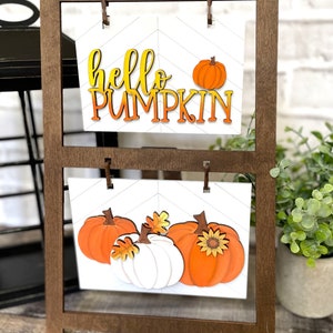 K&ouml;nnte beinhalten: Ein Holzst&auml;nder mit zwei Regalen, auf denen jeweils ein wei&szlig;es Schild mit K&uuml;rbis-Motiv steht. Das obere Schild zeigt den Text "hello pumpkin" in orangefarbenen und gelben Buchstaben. Das untere Schild zeigt drei K&uuml;rbisse, zwei orange und einen wei&szlig;en, mit Bl&auml;ttern und einer Sonnenblume.