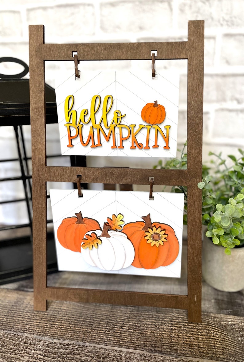 K&ouml;nnte beinhalten: Ein Holzst&auml;nder mit zwei Regalen, auf denen jeweils ein wei&szlig;es Schild mit K&uuml;rbis-Motiv steht. Das obere Schild zeigt den Text "hello pumpkin" in orangefarbenen und gelben Buchstaben. Das untere Schild zeigt drei K&uuml;rbisse, zwei orange und einen wei&szlig;en, mit Bl&auml;ttern und einer Sonnenblume.