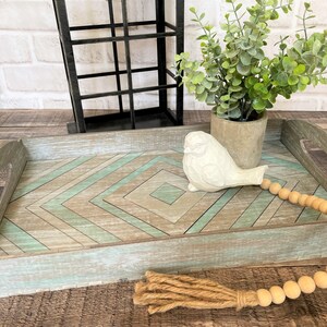 May include: A wooden tray with a geometric pattern in shades of brown and green. The tray features a white ceramic bird figurine and wooden beads.