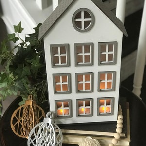May include: A white wooden house with nine windows and a gray roof. The house is decorated with two white and gold ornaments and a strand of wooden beads. The house is sitting on a stack of books.