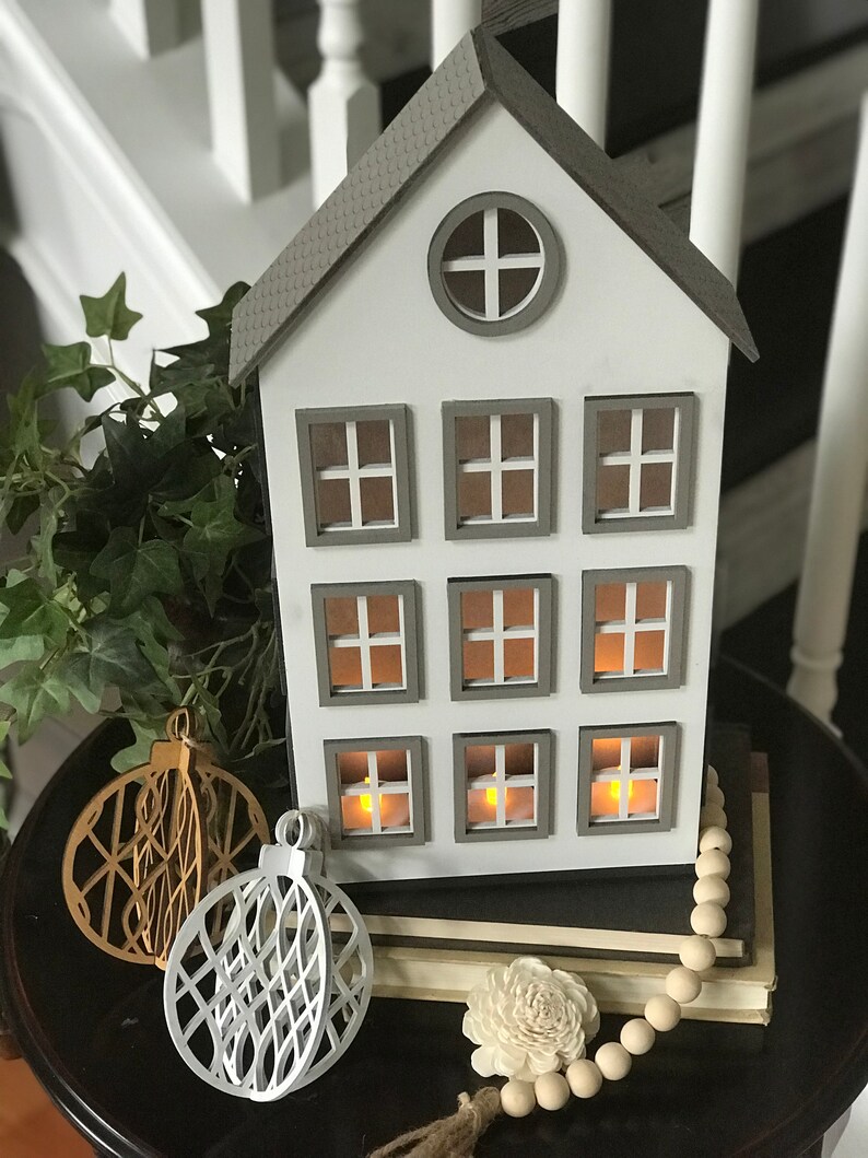 May include: A white wooden house with nine windows and a gray roof. The house is decorated with two white and gold ornaments and a strand of wooden beads. The house is sitting on a stack of books.