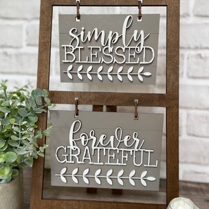 May include: A wooden easel with two signs. The top sign says "simply blessed" and the bottom sign says "forever grateful". Both signs have a gray background and white lettering.