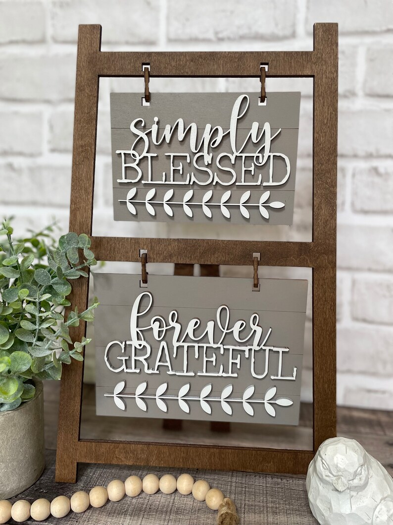 May include: A wooden easel with two signs. The top sign says "simply blessed" and the bottom sign says "forever grateful". Both signs have a gray background and white lettering.