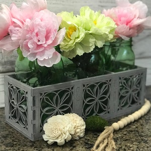 May include: A grey wooden planter with a decorative lattice design holds three glass jars filled with pink and green artificial flowers. A wooden bead garland with a jute tassel sits on a granite countertop.