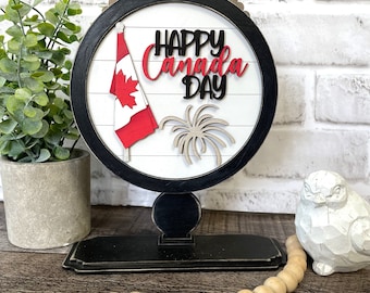 Puede incluir: Un letrero de madera blanco y negro con una bandera canadiense roja y blanca, fuegos artificiales y el texto "Feliz Día de Canadá". El letrero está en un soporte negro y está rodeado de cuentas de madera.