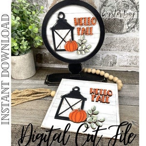 May include: A digital cut file design featuring a lantern, pumpkin, and leaves with the text "Hello Fall". The design is shown on a white wooden background with a black frame.