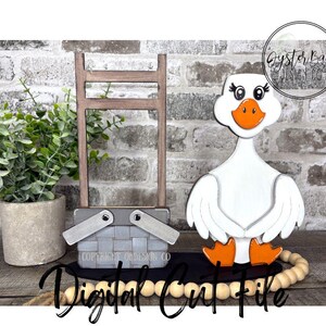 May include: A wooden goose figurine with an orange beak and feet, a picnic basket, and a small ladder are displayed on a black tray with wooden beads. A potted plant is to the left. The words "Digital Cut File" are written in cursive.