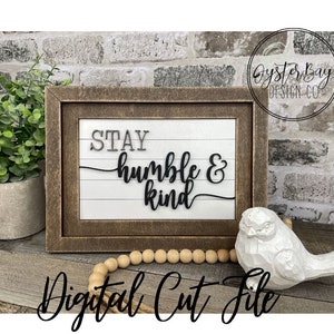 以下が含まれることがあります： 白い背景に黒い文字で「Stay humble & kind」と書かれた額縁付きの看板。看板は木製の表面に置かれ、白い鳥の置物と木製のビーズの紐が置かれている。