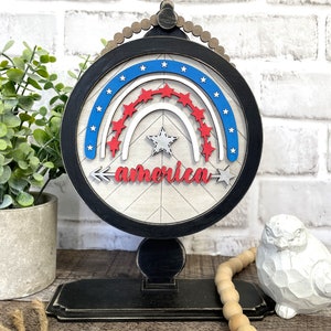 May include: A wooden sign with a red, white, and blue rainbow design and the word "america" in red lettering. The sign is on a black stand with a beaded trim.