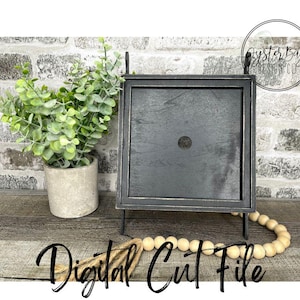 May include: Black square frame with a small hole in the centre, sitting on a wooden surface with a strand of wooden beads and a potted plant. The frame is on a black metal stand. The text "Digital Cut File" is written in black on the surface.