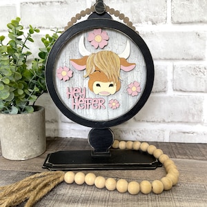 May include: A black wooden round sign with a white background and a painted brown and white cow head with pink flowers. The sign says "Hey Heifer" in pink letters. The sign is on a black stand with a wooden bead garland.