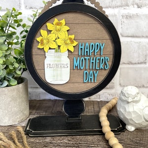 Può includere: Un cartello rotondo in legno con una cornice nera e un supporto. Il cartello ha uno sfondo in legno con assi dipinte di bianco. Il cartello dice "Happy Mother's Day" in lettere blu. C'è un arrangiamento di fiori gialli in un barattolo di vetro con la parola "Flowers" su di esso.