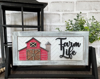 May include: A wooden sign featuring a red barn and silo illustration, with the text "Farm Life" in black script. The sign is displayed on a black easel with a wooden bead garland and a brown tassel.