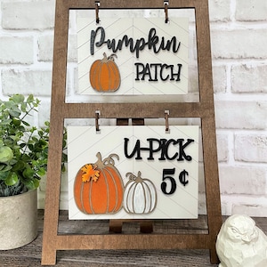 May include: A wooden easel sign with two signs. The top sign says "Pumpkin Patch" with a carved orange pumpkin. The bottom sign says "U-Pick 5¢" with two carved pumpkins, one orange and one white.