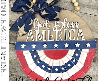 Puede incluir: Un letrero redondo de madera con el texto "God Bless America" en letras blancas. El letrero está decorado con un lazo azul y beige, un diseño en forma de abanico rojo, blanco y azul, y una cuerda de cuentas de madera.