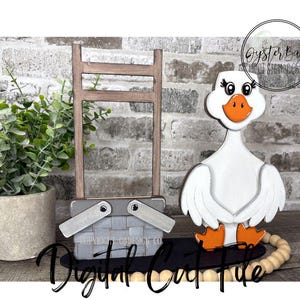 May include: A wooden goose figurine with orange feet and beak, white body, and a smiling face. A gray picnic basket and a wooden ladder are also present. The words "Digital Craft File" are written in cursive.