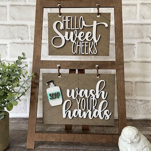 May include: A wooden bathroom sign holder with two signs. The top sign says "Hello Sweet Cheeks" and the bottom sign says "Wash Your Hands" with a small soap bottle graphic.