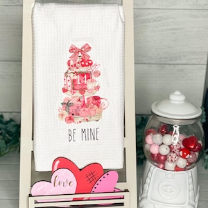 May include: White kitchen towel with a pink and red Valentine's Day themed design. The towel features a tiered cake with hearts, gifts, and the text "14 February". The towel also has the text "Be Mine". The towel is displayed on a white wooden stand with pink and red wooden hearts that say "love".