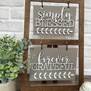 May include: A wooden easel with two signs. The top sign says "simply blessed" and the bottom sign says "forever grateful". Both signs have a white background with white lettering and a white leafy design at the bottom.