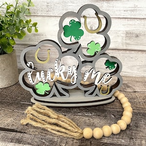 Può includere: Un cartello di legno con un design a strati di trifogli a quattro foglie e ferri di cavallo. Il cartello dice "lucky me" in un font bianco con puntini neri. Il cartello è decorato con trifogli verdi e ferri di cavallo dorati. Il cartello è su una superficie di legno con un fiocco marrone e perline di legno.