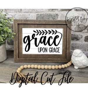 Pu&ograve; includere: Un cartello in legno incorniciato con sfondo bianco e lettere nere che recita "grace upon grace". Il cartello &egrave; decorato con un piccolo rametto di foglie nell'angolo in alto a sinistra. Il cartello &egrave; appoggiato su una superficie in legno con una collana di perline di legno e una statuetta di uccello in ceramica bianca.