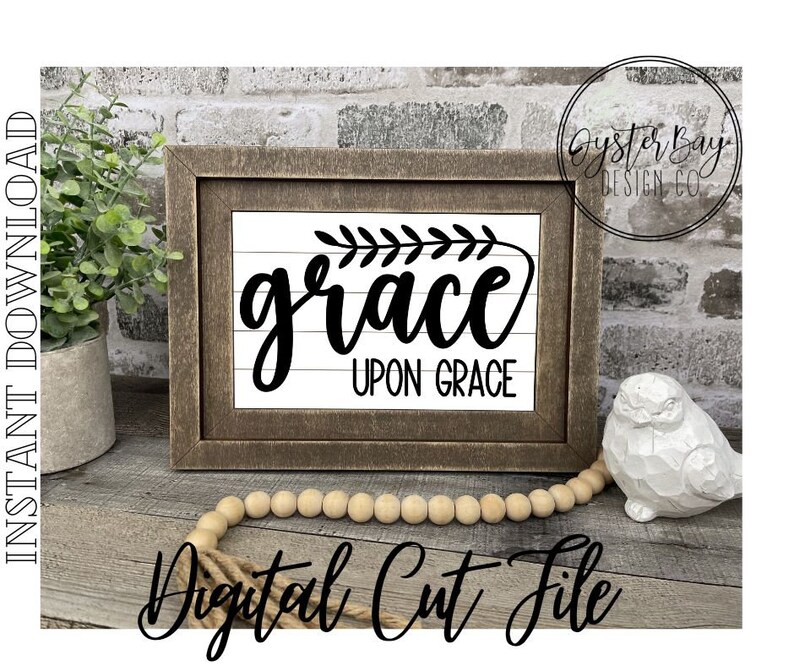 Pu&ograve; includere: Un cartello in legno incorniciato con sfondo bianco e lettere nere che recita "grace upon grace". Il cartello &egrave; decorato con un piccolo rametto di foglie nell'angolo in alto a sinistra. Il cartello &egrave; appoggiato su una superficie in legno con una collana di perline di legno e una statuetta di uccello in ceramica bianca.