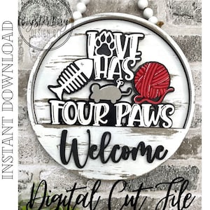 Op de afbeelding: Een rond, wit houten bord met de tekst "Love Has Four Paws Welcome" in zwarte letters. Het bord heeft een rode bol wol, een grijs katten speeltje, een visgraat en een pootafdruk.
