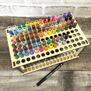 May include: A wooden stand with 100 holes for storing markers. The stand is made of light wood and has two tiers. The markers are arranged in a rainbow pattern. The stand is on a wooden surface.