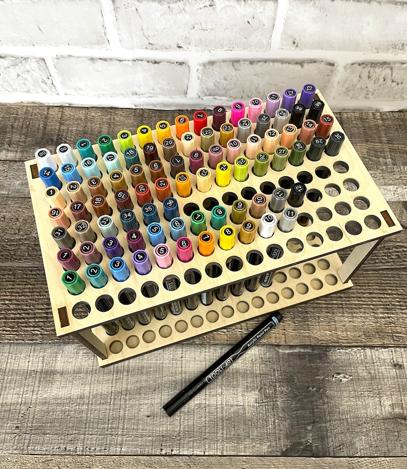May include: A wooden stand with 100 holes for storing markers. The stand is made of light wood and has two tiers. The markers are arranged in a rainbow pattern. The stand is on a wooden surface.