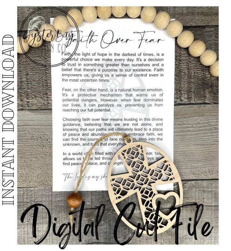 May include: A wooden cutout of a cross with a heart in the center. The cutout is surrounded by a decorative pattern. The cutout is hanging from a string with a wooden bead. The cutout is accompanied by a card with the text "Faith Over Fear" and a message about the importance of faith over fear.