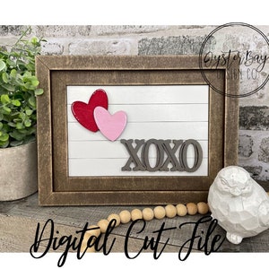 Puede incluir: Un letrero enmarcado de madera con fondo blanco y las palabras "XOXO" en letras negras. Hay dos corazones, uno rojo y otro rosa, en el letrero. El letrero está sobre una mesa con una guirnalda de cuentas de madera y una figurita de pájaro blanca.