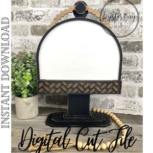 May include: A black wooden sign stand with a white sign insert and a decorative wood trim. The stand has a rounded top and a base with a woven design. The sign stand is on a wooden surface with a brick wall in the background. "Oyster Bay Design Co." is written on the sign stand.