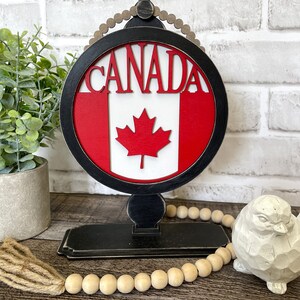 May include: A wooden Canada sign featuring a red and white Canadian flag and the word "CANADA" in red lettering. The sign is mounted on a black stand with a beaded trim. The sign is displayed on a wooden surface with a strand of wooden beads and a small white bird figurine.